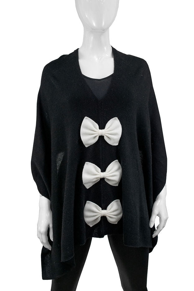 A black knit poncho sweater displayed on a white mannequin, featuring three large white bow details running vertically down the front.
