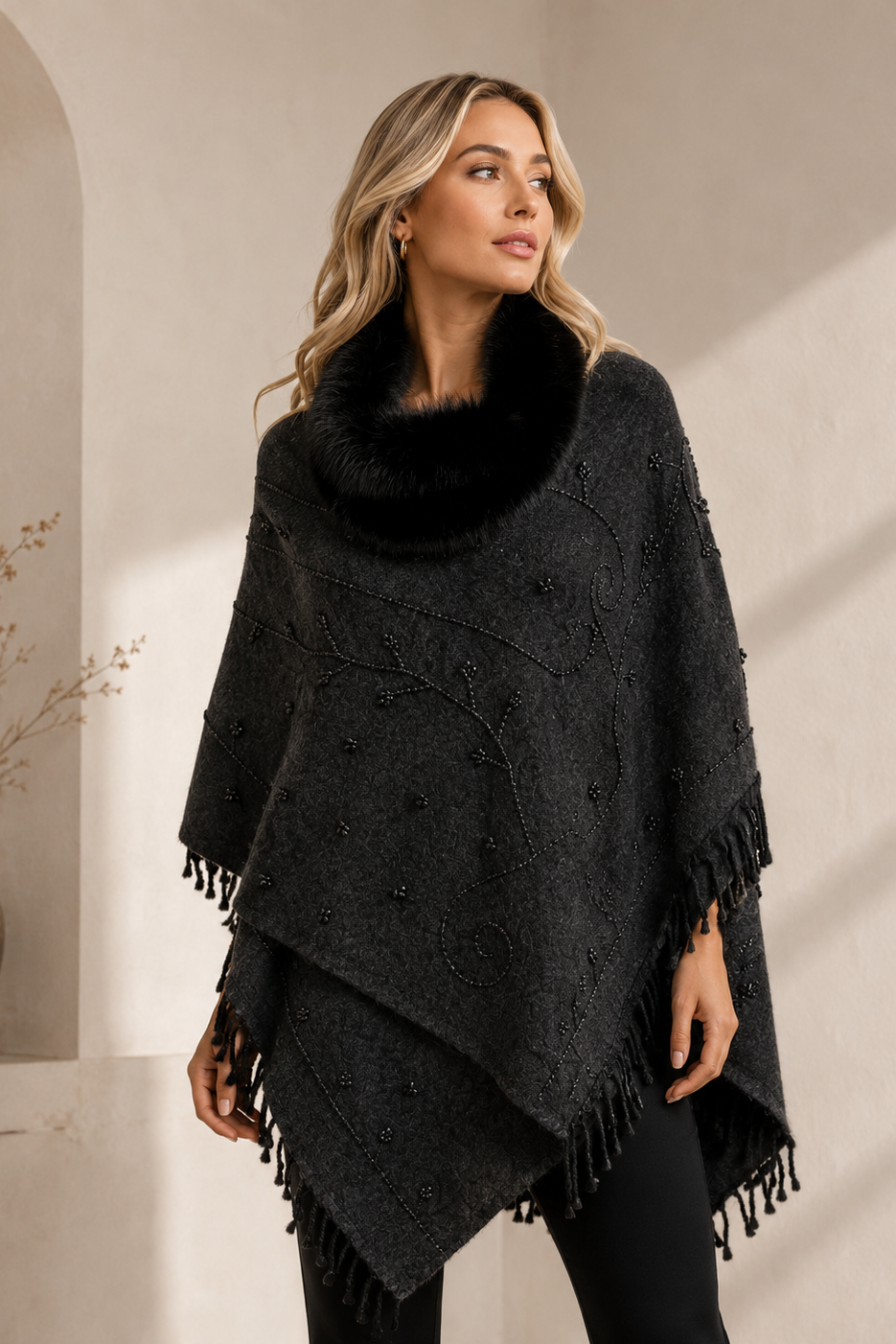 A woman wearing a black textured knit poncho with a black faux-fur collar and fringe trim, standing in a warm, sunlit interior setting.