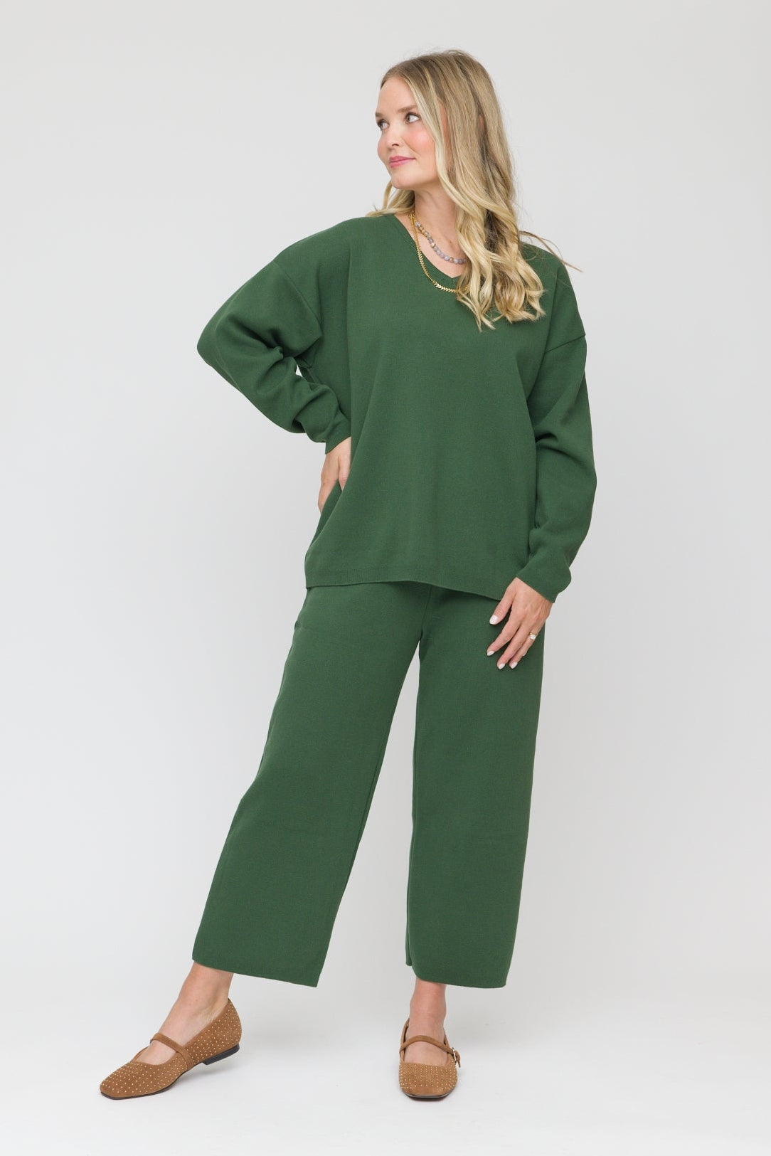 A woman modeling a matching dark green lounge set consisting of a long-sleeve V-neck top and wide-leg cropped pants, paired with tan flats, against a plain light gray studio background.