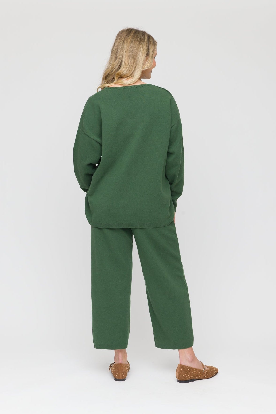 A woman viewed from behind wearing a matching dark green lounge set consisting of a long-sleeve top and wide-leg cropped pants, paired with brown flat shoes, against a light gray studio background.