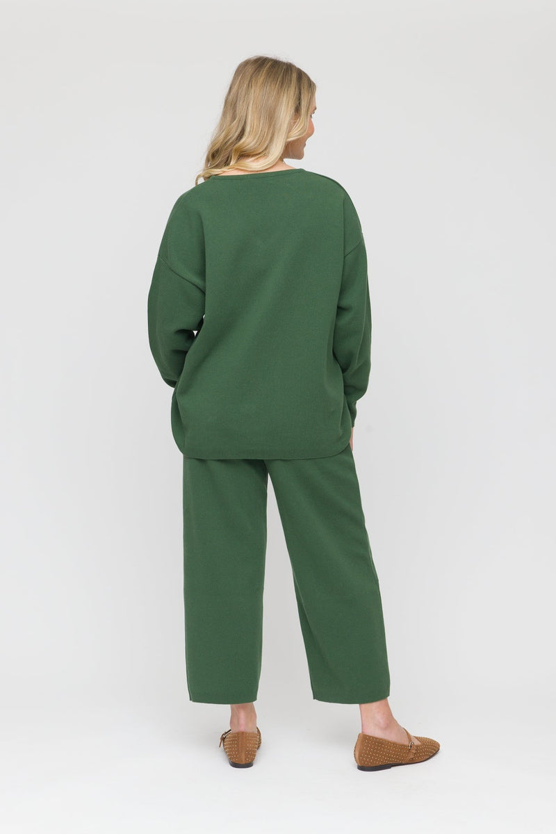 A woman viewed from behind wearing a matching dark green lounge set consisting of a long-sleeve top and wide-leg cropped pants, paired with brown flat shoes, against a light gray studio background.