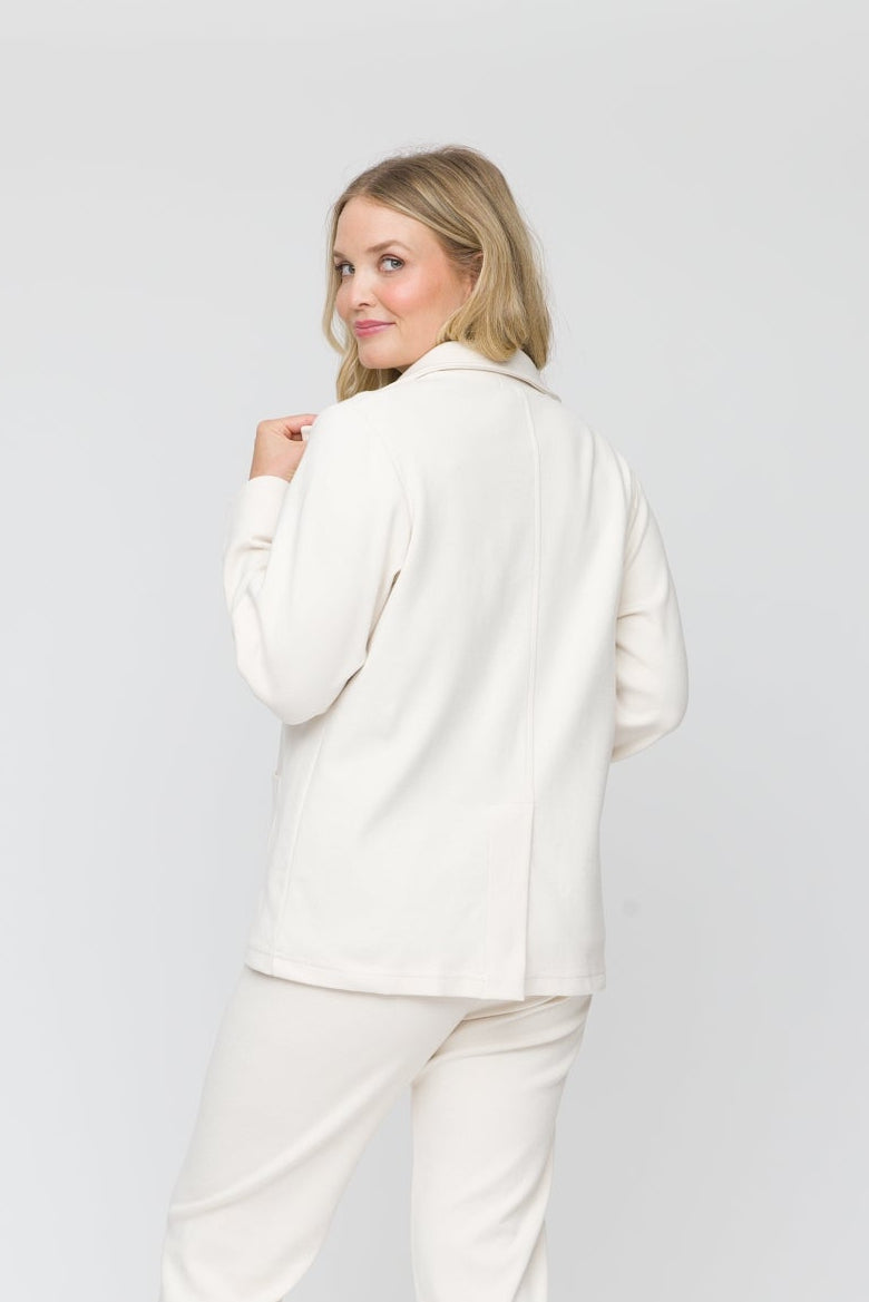 A woman modeling a matching cream-colored two-piece set consisting of a long-sleeve button-up shirt and trousers, shown from a back-three-quarter view against a white studio backdrop.