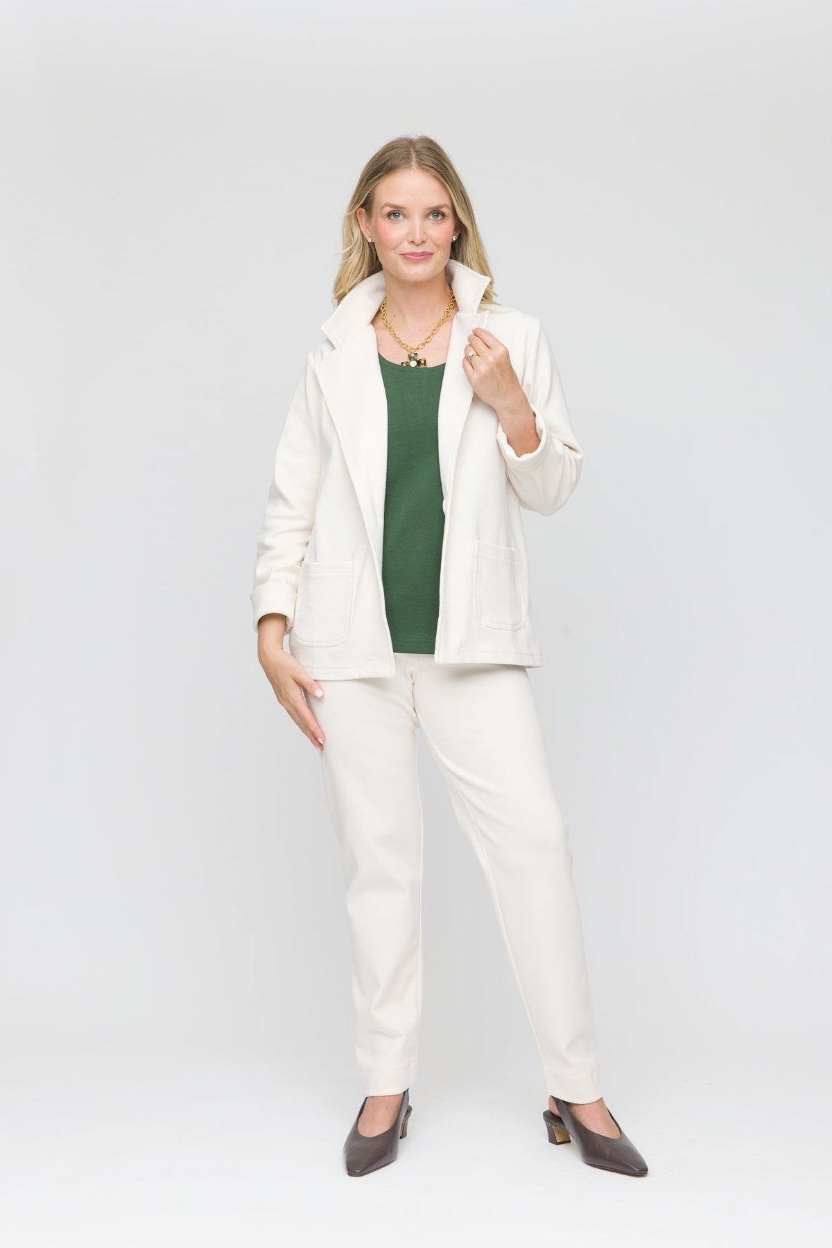 A woman modeling a white two-piece blazer and trouser set over a dark green top, paired with dark brown pointed-toe heels, against a seamless white studio background.